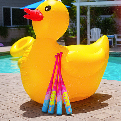 A large yellow inflatable duck pool float sits on a patio by a pool. Three Easton 2025 Hype Fire Pool Party (-5) 2 3/4" USSSA baseball bats in pink and blue are propped against the duck.