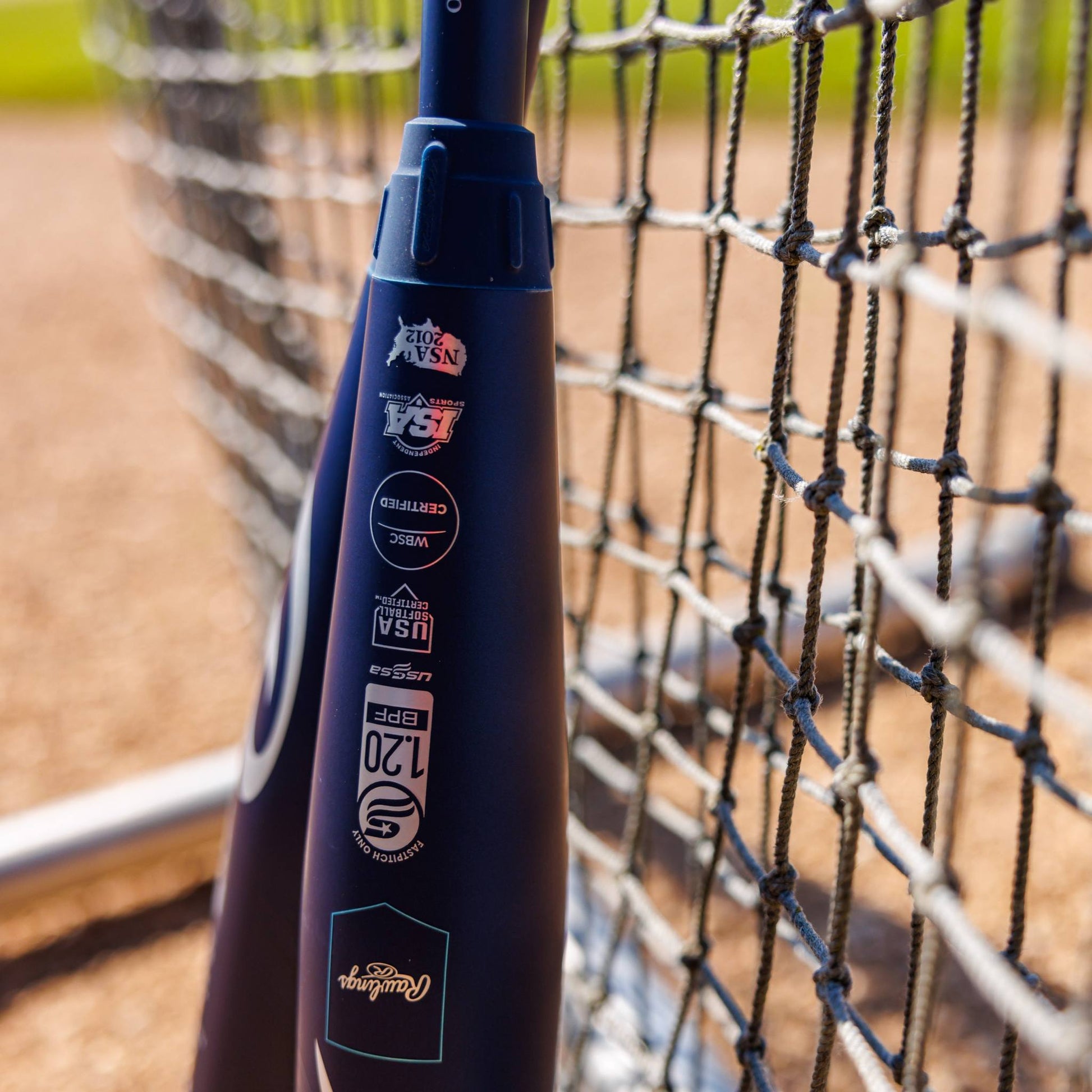 A close-up of the 2025 Rawlings Mantra (-10) Fastpitch Softball Bat: RFP4M10 (DEMO) by Rawlings leaning on a chain-link fence, displaying certification stickers with a blurred softball field in the background.