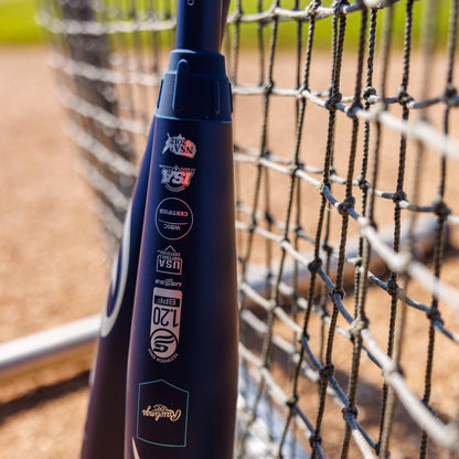 A close-up of the 2025 Rawlings Mantra (-10) Fastpitch Softball Bat: RFP4M10 (DEMO) by Rawlings leaning on a chain-link fence, displaying certification stickers with a blurred softball field in the background.
