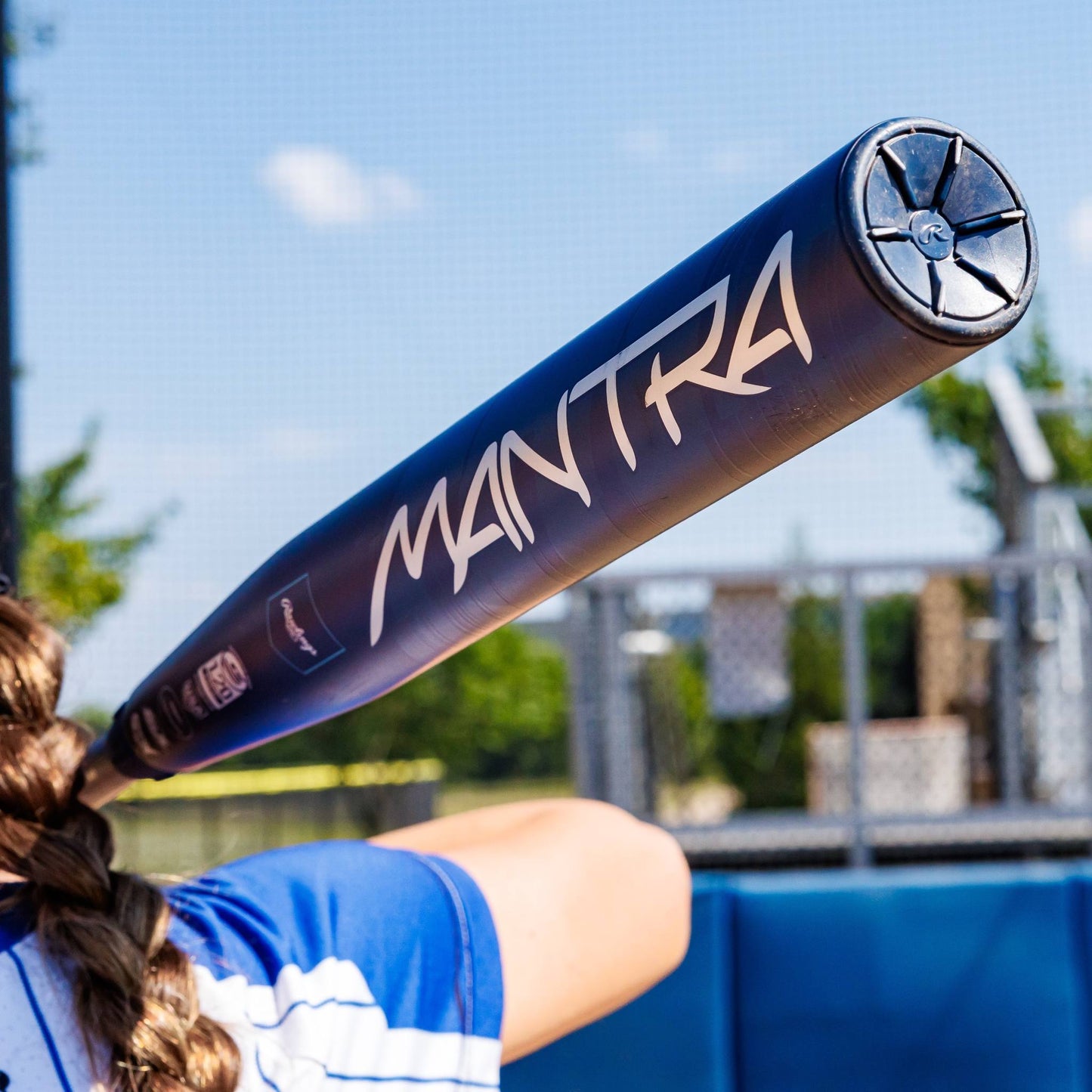 A person in a blue and white jersey holds up a 2025 Rawlings Mantra (-10) Fastpitch Softball Bat: RFP4M10 (DEMO) as “MANTRA” stands out, with a blurred outdoor fastpitch softball field and fence in the background.