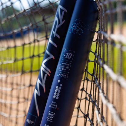 Two Rawlings 2025 Mantra (-10) Fastpitch Softball Bats (RFP4M10 DEMO) lean against a net fence on the field, green grass and dirt blurred in the background—a top choice for players seeking premium Rawlings performance.