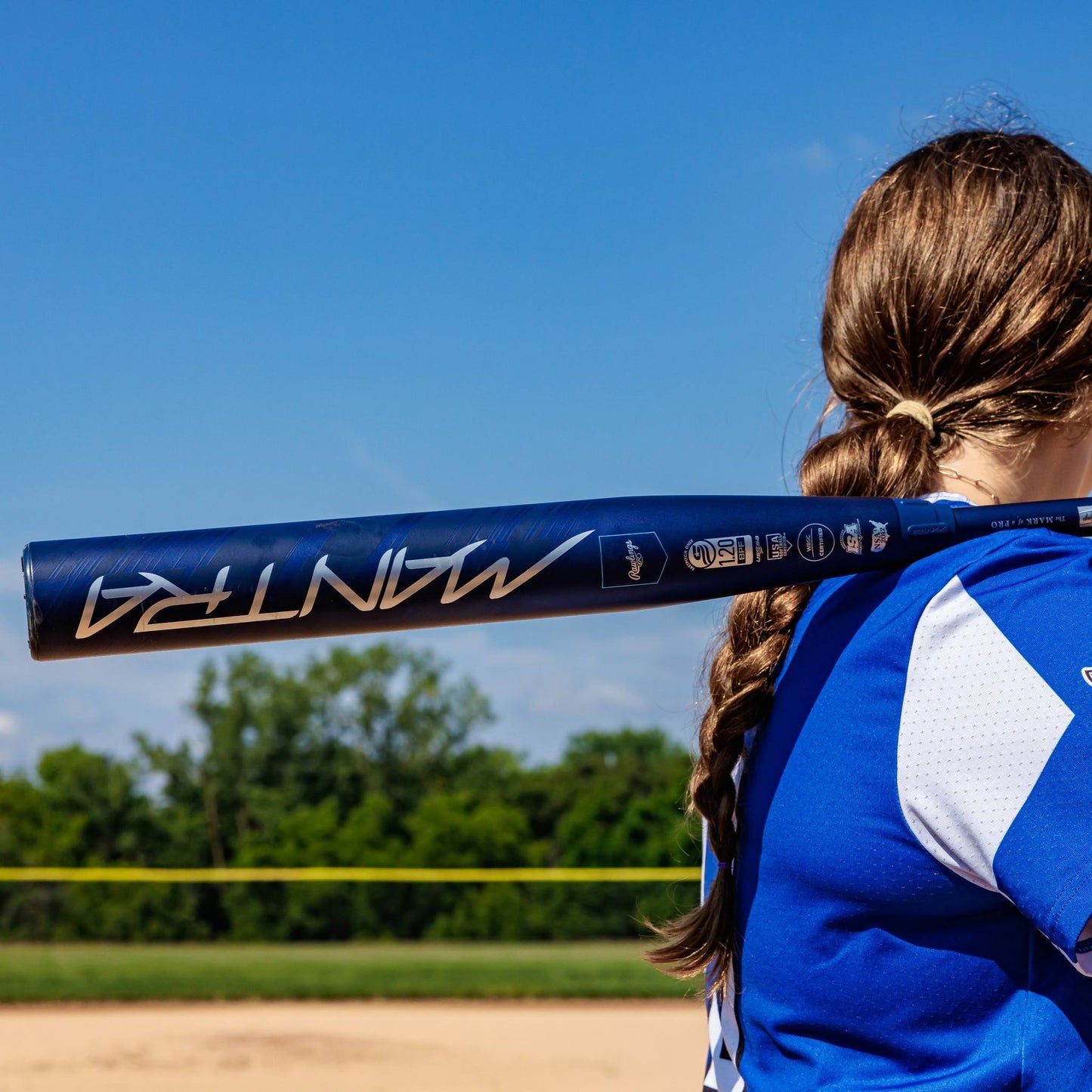 A softball player with long brown hair in a braid stands on a sunny field, holding the 2025 Rawlings Mantra (-10) Fastpitch Softball Bat (RFP4M10 DEMO) on her shoulder, surrounded by green trees and a clear blue sky.