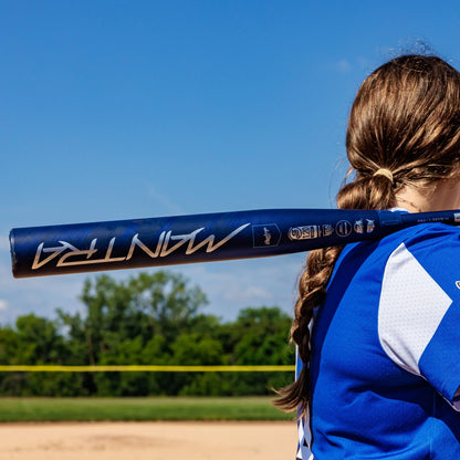 A softball player with long brown hair in a braid stands on a sunny field, holding the 2025 Rawlings Mantra (-10) Fastpitch Softball Bat (RFP4M10 DEMO) on her shoulder, surrounded by green trees and a clear blue sky.