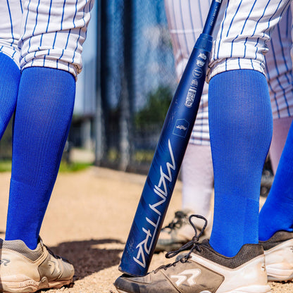 Several athletes in blue knee-high socks, white pinstripe pants, and cleats stand on a dirt field. A 2025 Rawlings Mantra (-10) Fastpitch Softball Bat: RFP4M10 (DEMO) from Rawlings leans against one player’s leg.