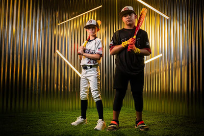 Two young boys in baseball uniforms stand on green turf, confidently holding the Easton 2025 Hype Fire (-8) 2 5/8" USA Baseball Bat, with a metallic gold-lit background and geometric neon lights behind them.