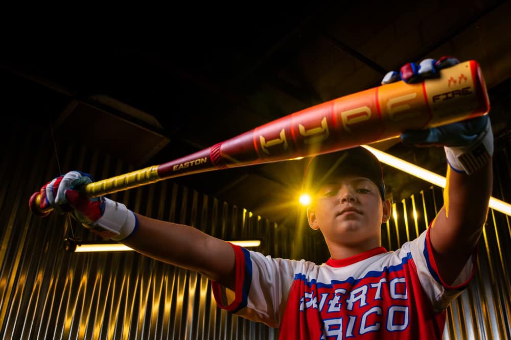A young baseball player in a Puerto Rico jersey holds an Easton 2025 Hype Fire (-8) 2 5/8" USA Baseball Bat (EUS5HYP8), posed facing the camera with dramatic lighting and corrugated metal walls setting a focused mood.