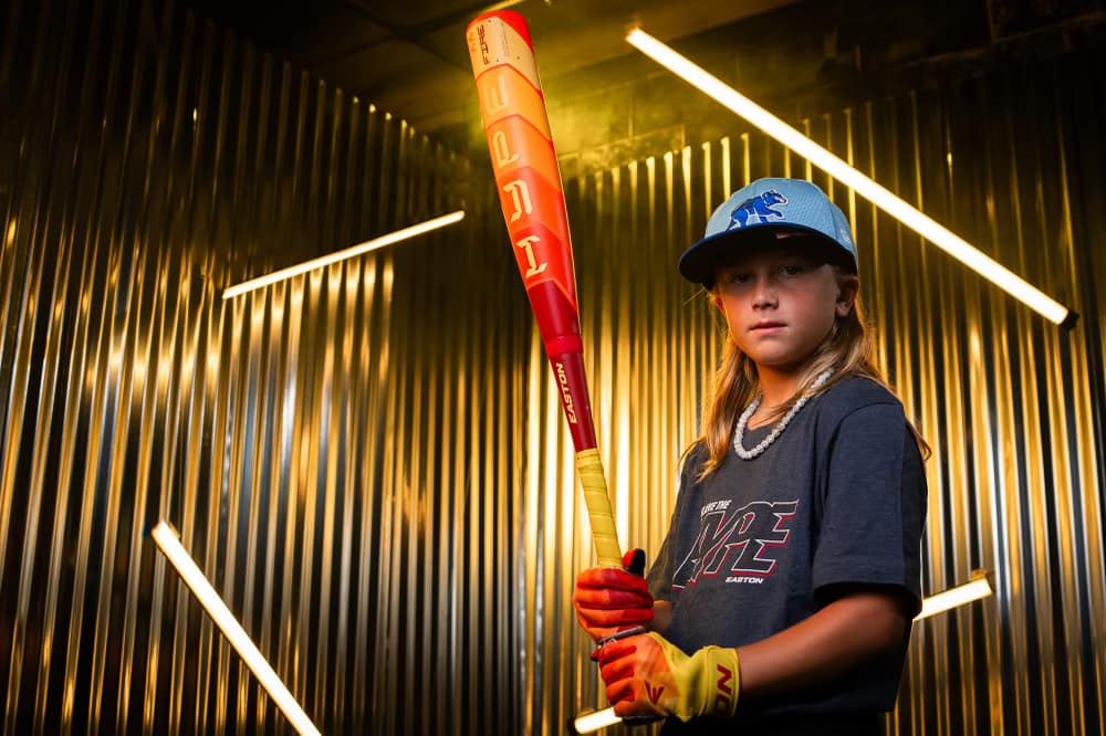 A young baseball player in a blue hat and gray shirt holds the 2025 Easton Hype Fire (-8) 2 5/8" USA Baseball Bat (EUS5HYP8) by Easton, wearing yellow gloves and standing in front of metallic corrugated walls lit by diagonal lights.