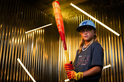 A young baseball player in a blue hat and gray shirt holds the 2025 Easton Hype Fire (-8) 2 5/8" USA Baseball Bat (EUS5HYP8) by Easton, wearing yellow gloves and standing in front of metallic corrugated walls lit by diagonal lights.