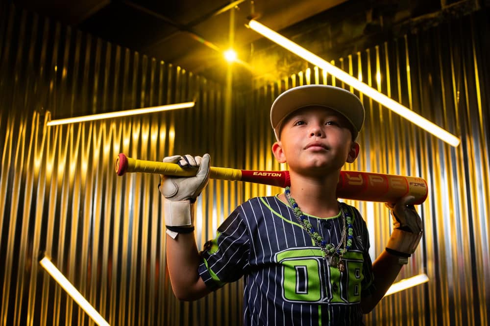 A young boy in a baseball uniform and cap stands confidently with the 2025 Easton Hype Fire (-8) 2 5/8" USA Baseball Bat (EUS5HYP8) from Easton across his shoulders, framed by metallic walls and glowing light tubes.