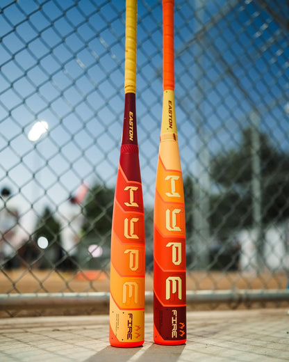 Two colorful baseball bats, including the 2025 Easton Hype Fire (-5) 2 5/8" USA Baseball Bat (EUS5HYP5) by Easton, stand upright against a chain-link fence on a baseball field with blurred trees and sky in the background.