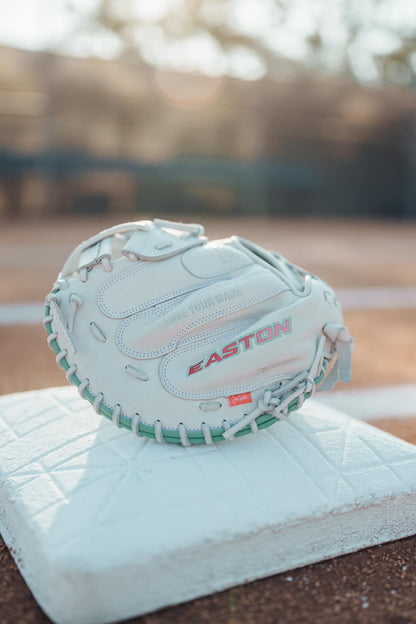 The Easton Jen Schro "The Rudi" 34" Fastpitch Catcher's Mitt (RUDI-2024), made of premium USA steerhide, sits on a white base on a baseball field with sunlight in the background.