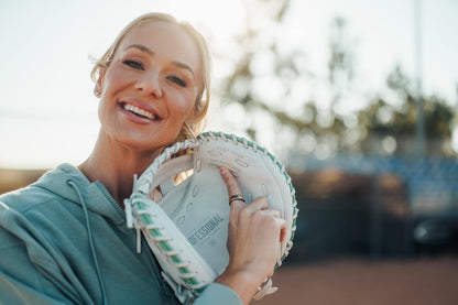 A smiling woman in a light green hoodie holds the Easton Jen Schro "The Rudi" 34" Fastpitch Catcher's Mitt (RUDI-2024) and points to text inside it. She stands outdoors on a sunny day with blurred trees and a field in the background.