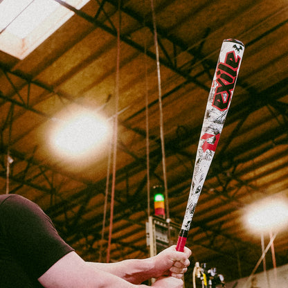 A person grips a DeMarini 2026 Exile (-3) BBCOR Baseball Bat (WBD2589010) with both hands, preparing to swing inside an indoor facility featuring a high wooden ceiling and exposed beams.