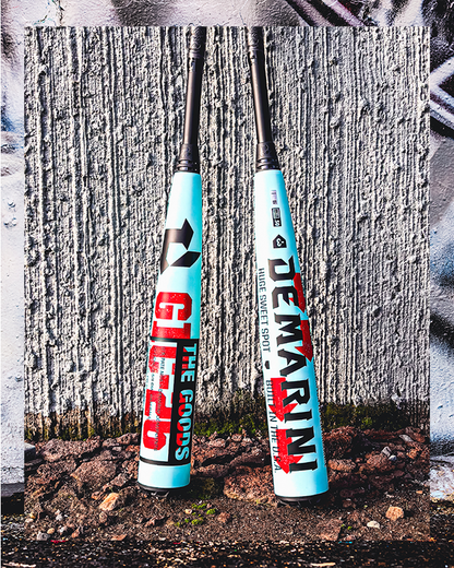 Two 2026 DeMarini The Goods (-3) BBCOR Baseball Bats (WBD2588010) with bold red and black graphics stand upright against a graffiti-covered concrete wall, with dirt and small rocks at the base of each DeMarini hybrid bat.