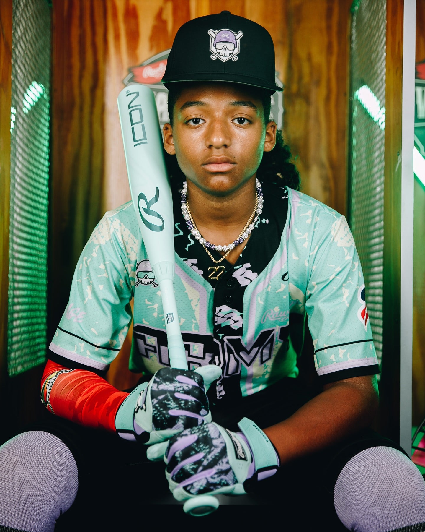 A young baseball player in a mint green and white jersey holds the 2026 Rawlings Icon (-5) 2 3/4" USSSA Baseball Bat: RUT6I5. He wears a black cap, gloves, and red arm sleeve, looking at the camera with a serious expression.