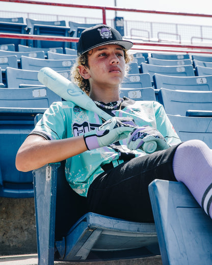 A young baseball player in a teal jersey and cap sits in stadium seats, gripping a Rawlings 2026 Icon (-5) 2 3/4" USSSA Baseball Bat (RUT6I5) and wearing batting gloves, gazing thoughtfully into the distance.