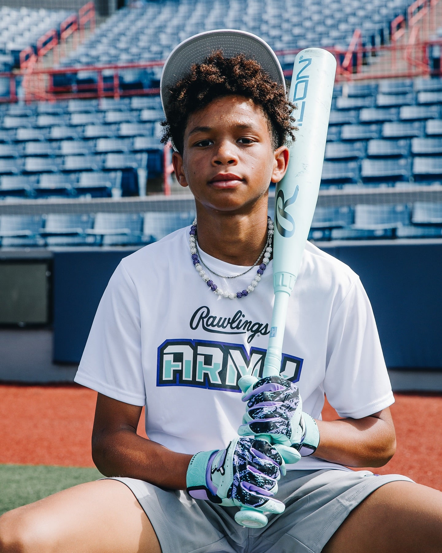 A young baseball player sits in empty stadium seats, gripping the Rawlings Icon (-5) 2 3/4" USSSA Baseball Bat (RUT6I5) and wearing a white Rawlings Army shirt, patterned batting gloves, and a backward cap.