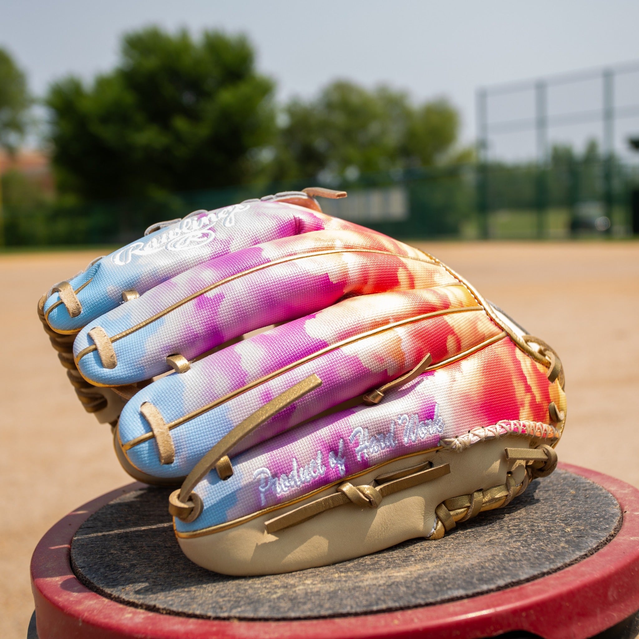 A multicolored baseball glove with blue, pink, orange, and yellow patterns sits on a red surface at a baseball field, with green trees and a fence visible in the background.