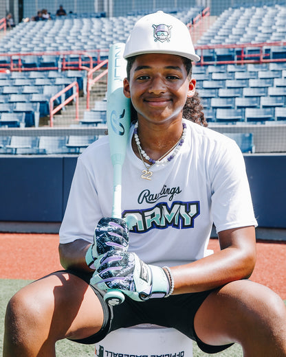 A young baseball player in a white Rawlings Army shirt, cap, and gloves sits on a bench holding the 2026 Rawlings Icon (-5) 2 3/4" USSSA Baseball Bat (RUT6I5), smiling at the camera in an empty stadium.