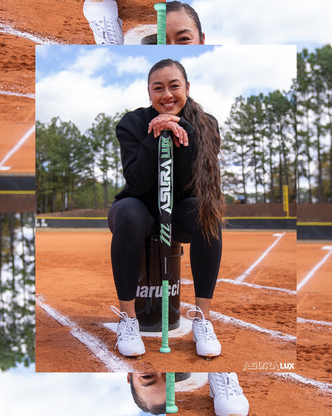 A smiling woman in athletic wear sits on a bucket on a softball field, holding the Marucci 2026 ASURA Lux (-11) Fastpitch Softball Bat (MFPASL11). Trees, blue sky, infield dirt, and bases are visible in the background.