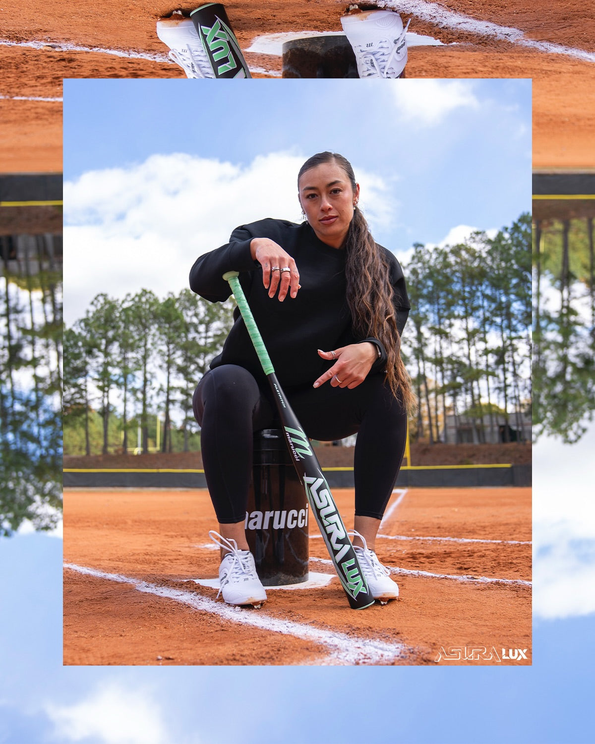 A woman in black athletic wear sits on a bucket on a softball field, holding the 2026 Marucci ASURA Lux (-11) Fastpitch Softball Bat (MFPASL11). Pine trees and a blue sky with clouds appear, framed by a stylized border.