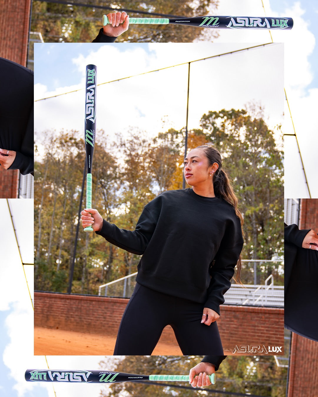 A woman in a black sweatshirt and leggings stands on a baseball field, holding the Marucci 2026 ASURA Lux (-10) Fastpitch Softball Bat (MFPASL10) vertically with one hand. Autumn trees and a chain-link fence are visible in the background.