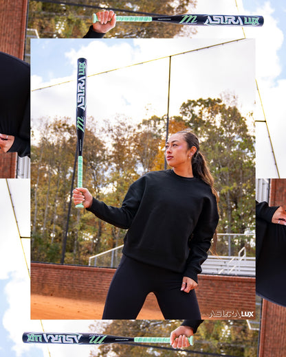 A woman in a black sweatshirt and leggings stands on a baseball field, holding the Marucci 2026 ASURA Lux (-10) Fastpitch Softball Bat (MFPASL10) vertically with one hand. Autumn trees and a chain-link fence are visible in the background.