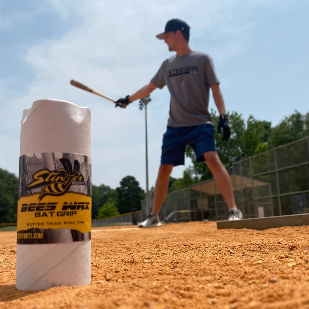 A Stinger BEES WAX Bat Grip by Stinger Sports sits near home plate on a sunny baseball field, as a player prepares to bat. This beeswax grip provides an effective pine tar alternative for better hold.