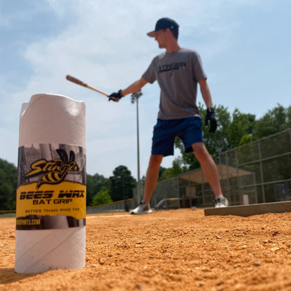 A Stinger BEES WAX Bat Grip by Stinger Sports sits near home plate on a sunny baseball field, as a player prepares to bat. This beeswax grip provides an effective pine tar alternative for better hold.