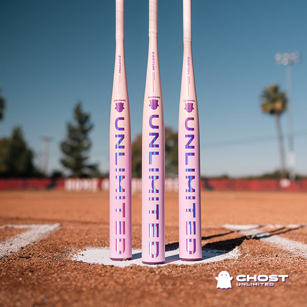 Three white and purple 2026 Easton Ghost Unlimited (-10) Fastpitch Softball Bats (EFP6GHUL10) stand upright on a softball field, with trees and a scoreboard blurred behind them. The Easton Ghost Unlimited Double Barrel EXT logo appears in the lower right corner.