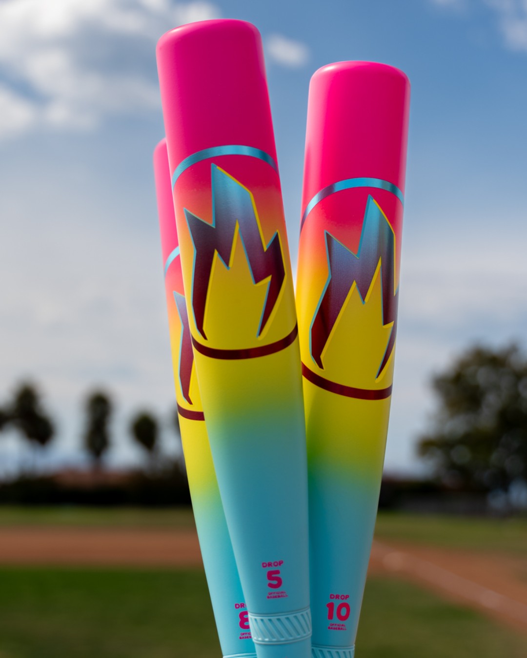 Three eye-catching 2026 Easton Hype Fire (-5) USSSA Baseball Bats, featuring a pink, yellow, and turquoise gradient with a flame logo, are shown upright outdoors with a blurred baseball field and trees in the background.