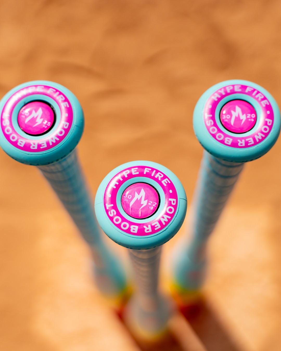 Three Easton Hype Fire (-5) USSSA Baseball Bats (2026) with blue handles and pink HYPE FIRE POWER BOOST end caps stand upright on sand, showing the flame icon, weight marks, and ThermoComposite tech from above.