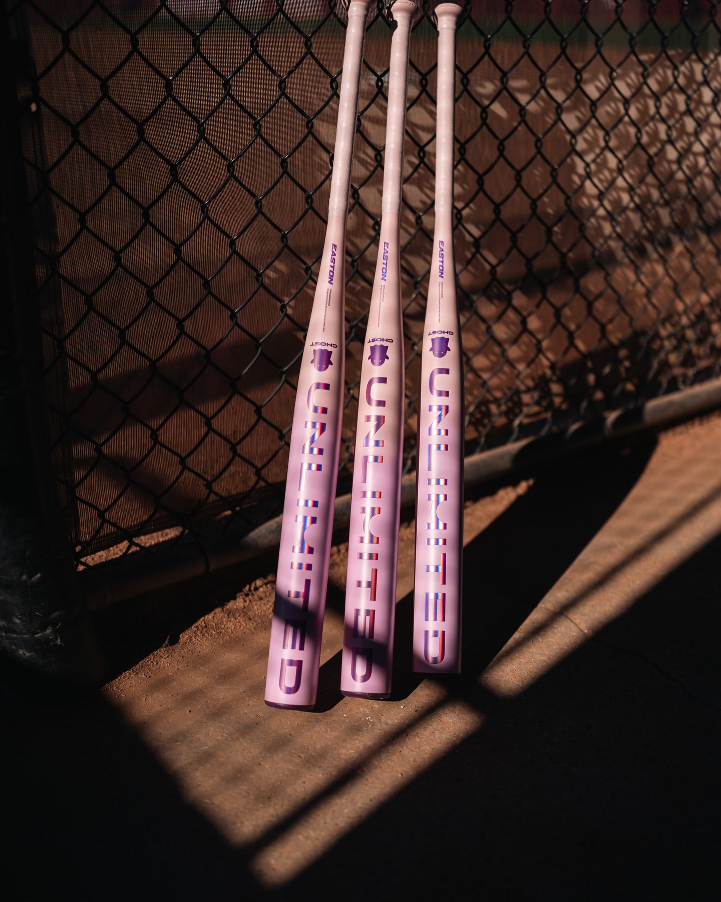 Three light purple Easton Ghost Unlimited (-8) Fastpitch Softball Bats (EFP6GHUL8) with "UNLIMITED" in bold letters lean against a chain-link fence, sunlight casting partial shadows on the ground.