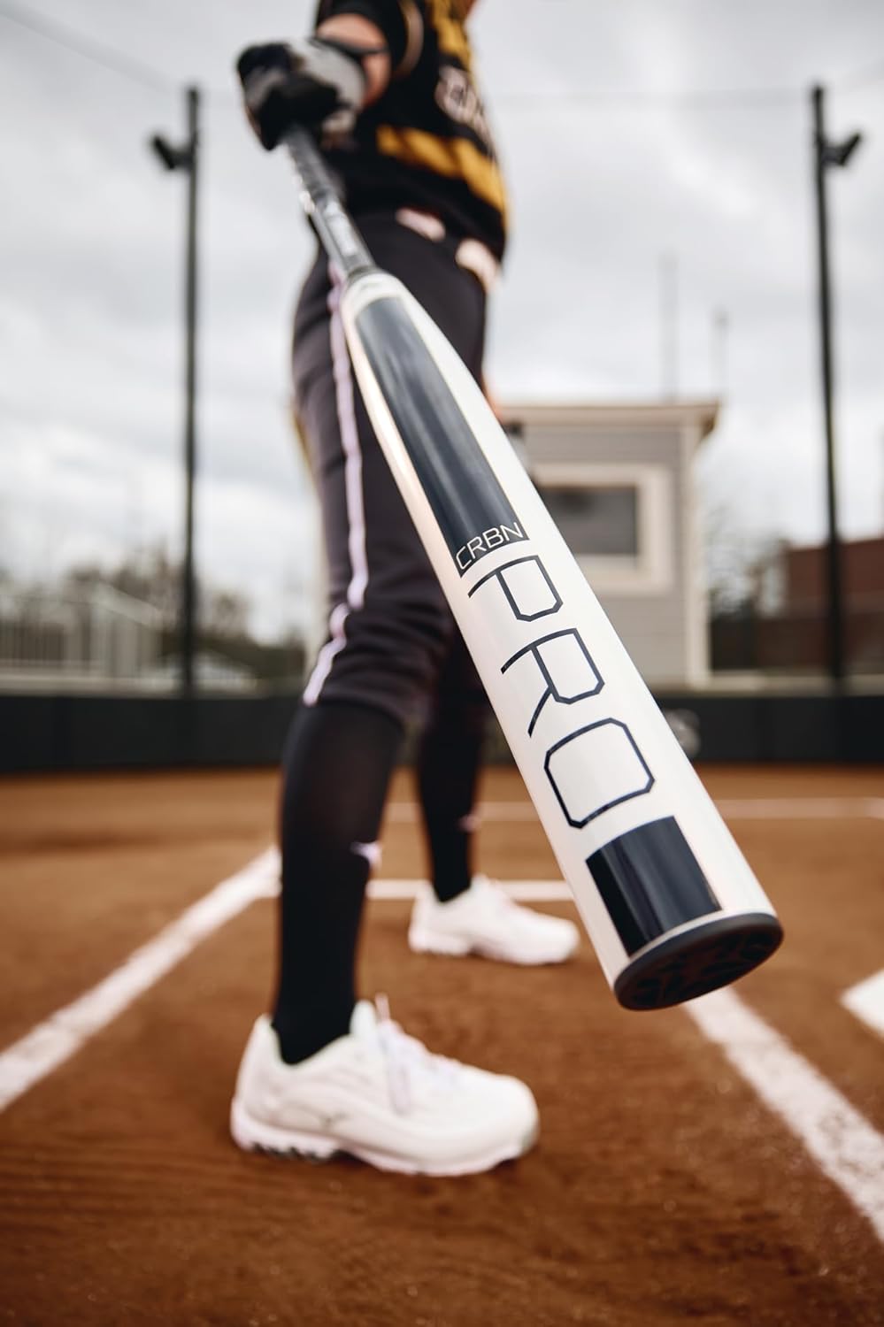 A baseball player in black pants and white shoes holds a Mizuno 2025 CRBN Pro (-10) Fastpitch Softball Bat (340665) on a field, with a small building and field fences visible in the background.