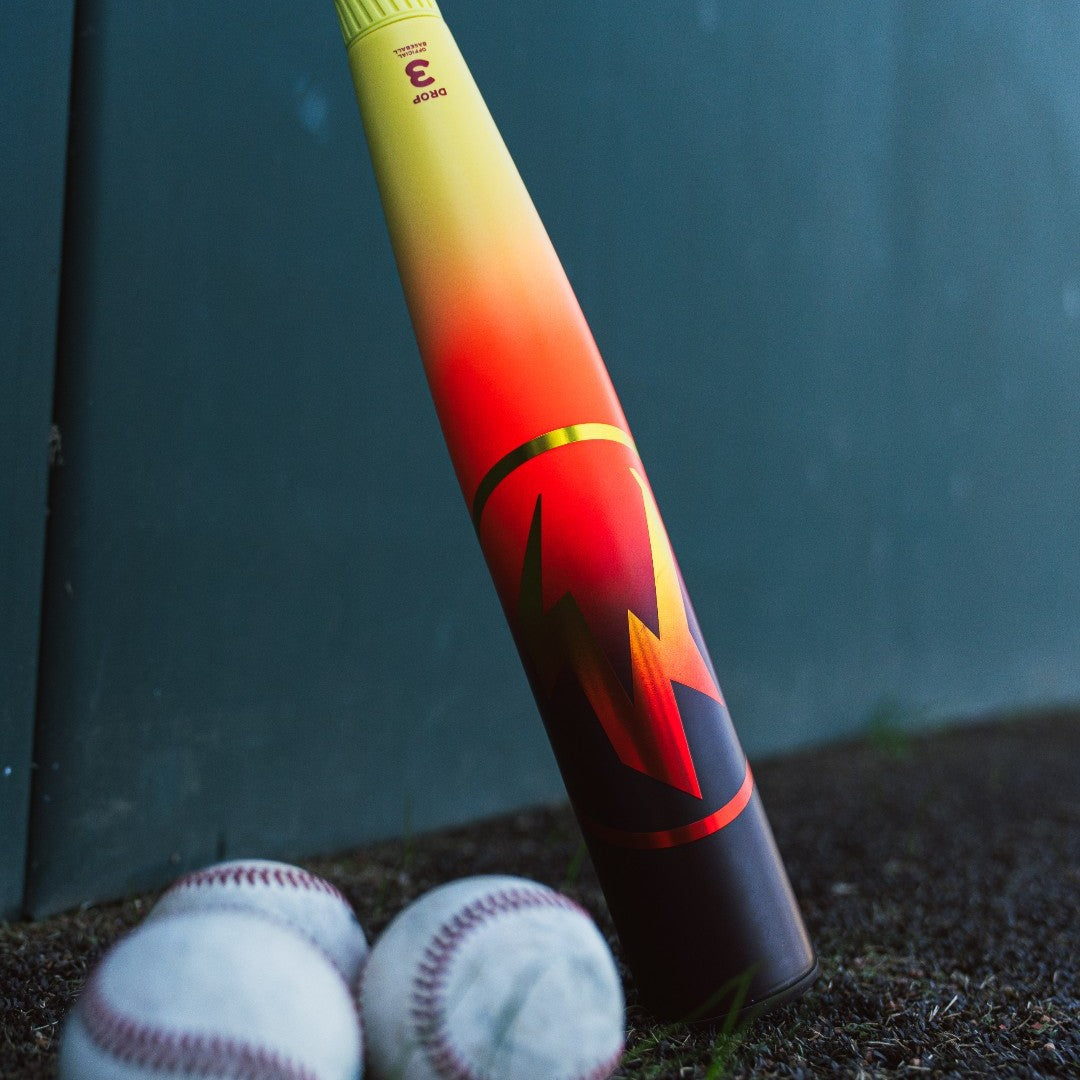 The 2026 Easton Hype Fire (-3) BBCOR Baseball Bat by Easton features a red-yellow gradient and bold lightning design, standing upright near three white baseballs against a green wall background.