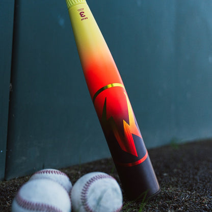 The 2026 Easton Hype Fire (-3) BBCOR Baseball Bat by Easton features a red-yellow gradient and bold lightning design, standing upright near three white baseballs against a green wall background.