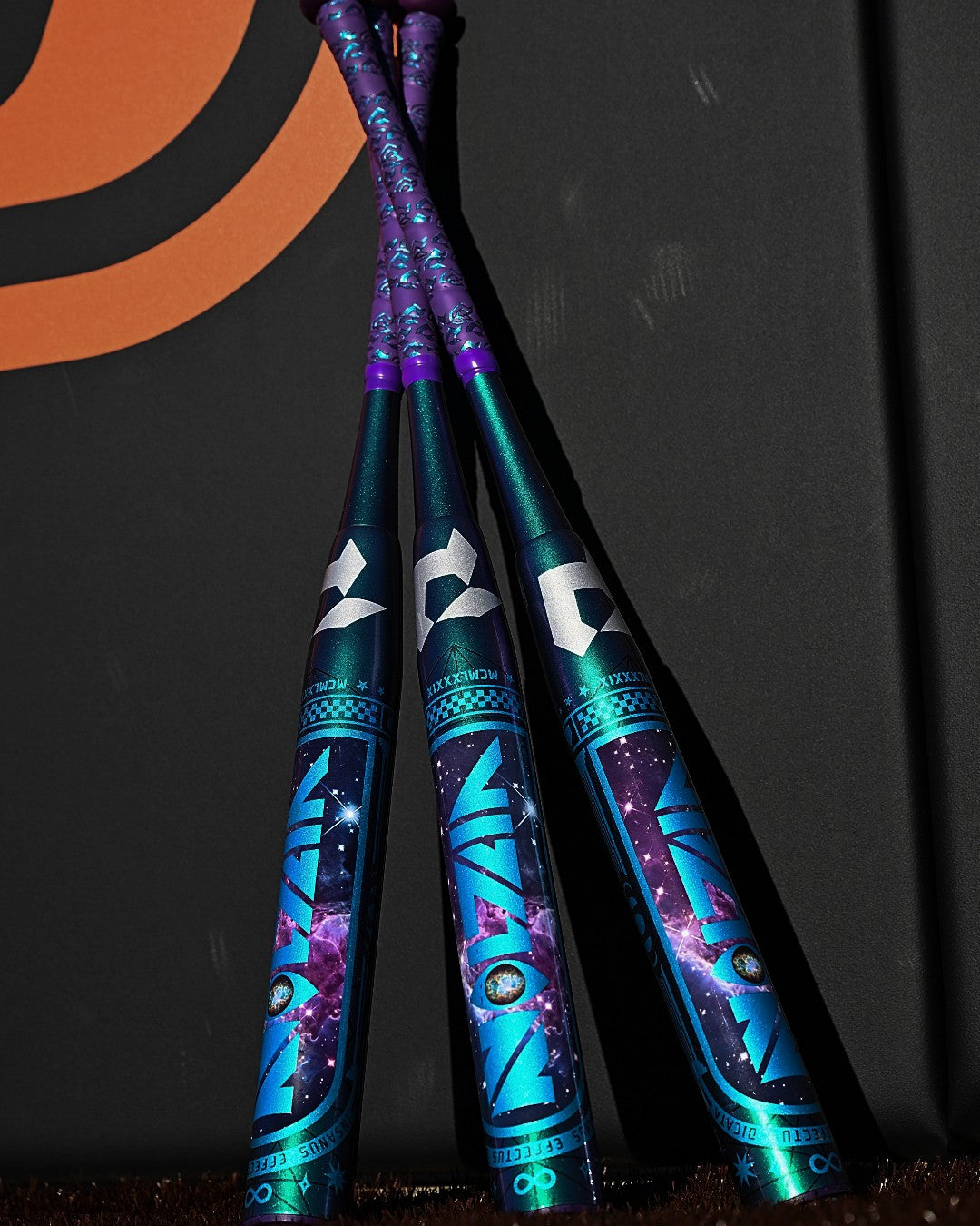 Three metallic 2027 DeMarini Vizion (-8) Fastpitch Softball Bats (WBD2730010), featuring blue and purple cosmic designs, stand upright crossed at the top, leaning against a black wall with an orange swirl logo.