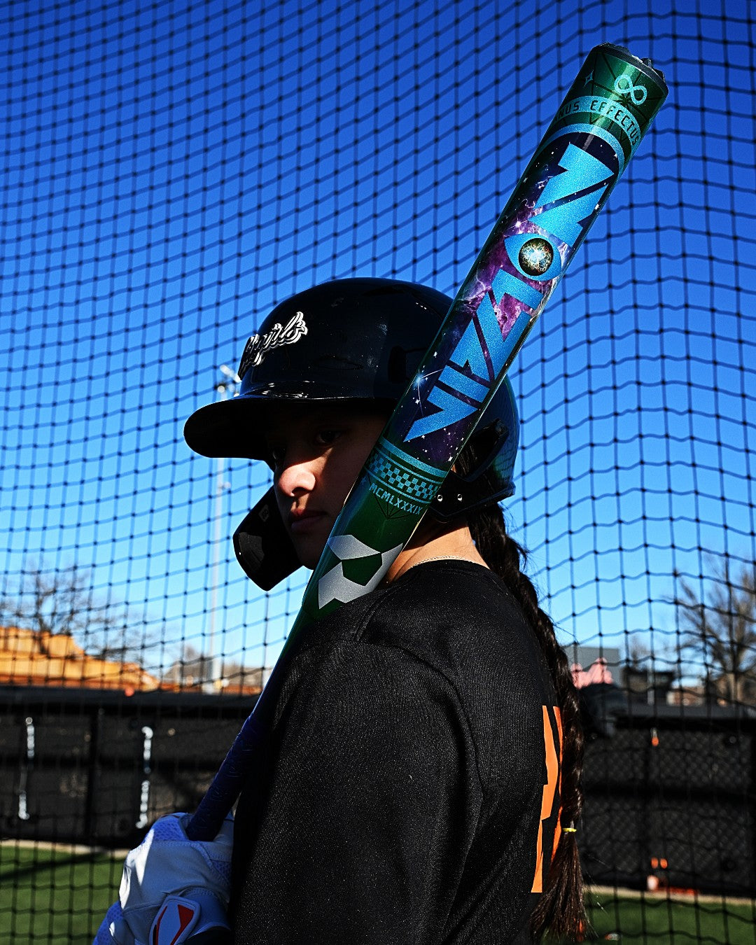 A softball player in black gear stands on a field with trees and a net fence, resting the DeMarini 2027 Vizion (-8) Fastpitch Softball Bat (WBD2730010) on her shoulder.
