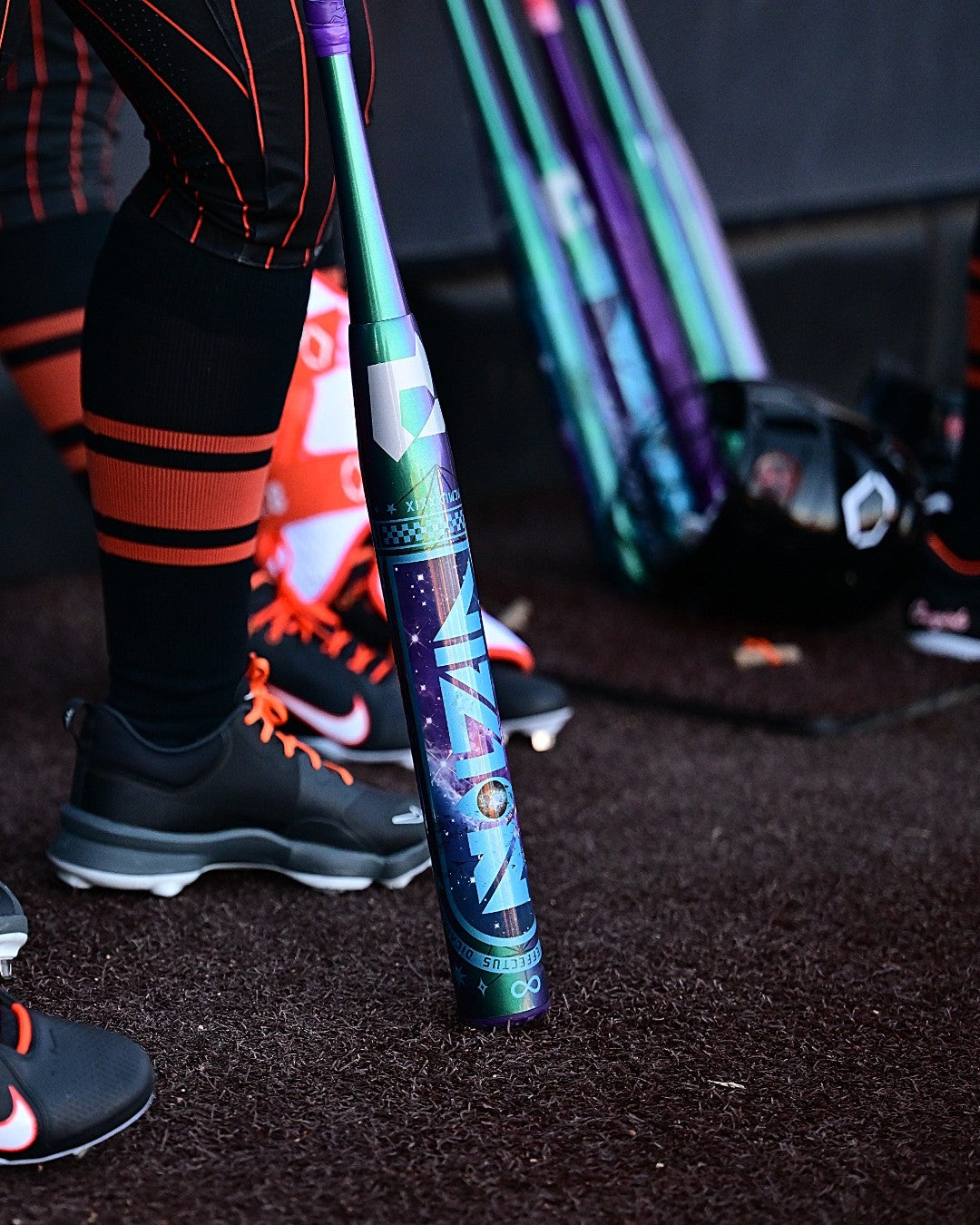 A person in black and orange athletic gear stands beside a DeMarini Vizion 2027 (-8) Fastpitch Softball Bat (WBD2730010) on dark turf, with more DeMarini composite bats, a helmet, and other gear visible in the background.