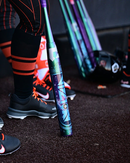A person in black and orange athletic gear stands beside a DeMarini Vizion 2027 (-8) Fastpitch Softball Bat (WBD2730010) on dark turf, with more DeMarini composite bats, a helmet, and other gear visible in the background.