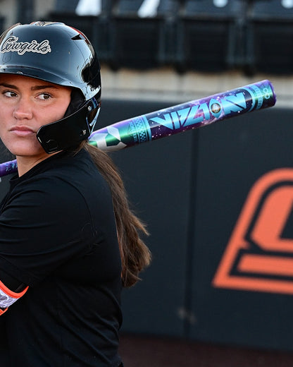 A softball player in a black helmet and uniform rests a DeMarini 2027 Vizion (-8) Fastpitch Softball Bat (WBD2730010) on her shoulder, glancing sideways. Behind her are empty stadium seats and a wall with an orange and black logo.
