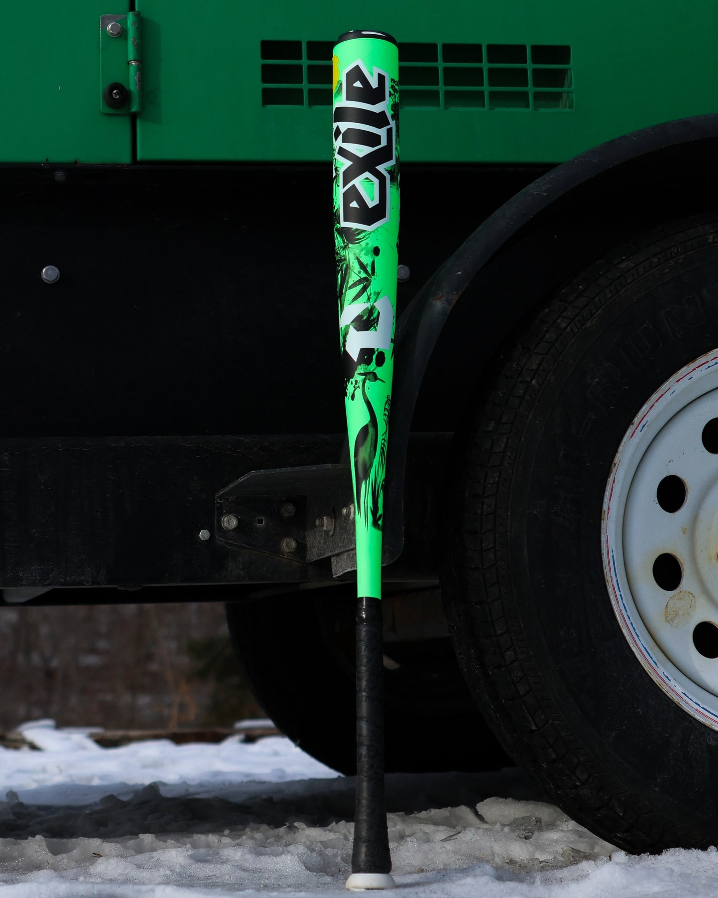 A 2026 DeMarini Exile Samurai (-3) BBCOR Baseball Bat (WBD2652010) with bright green and black design leans upright against the wheel of a large black vehicle on snowy ground.