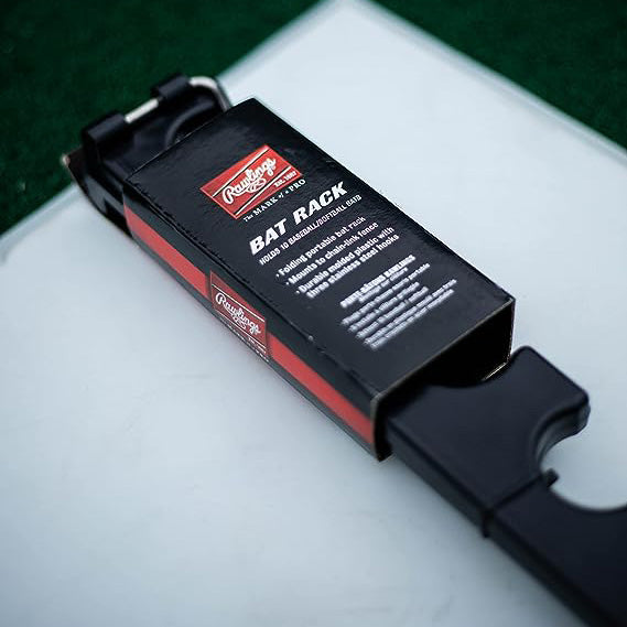 A Rawlings Portable Fence Bat Rack (BATRACK), in black packaging, rests on a white surface with green turf in the background. The box features the Rawlings brand and product name in white text.