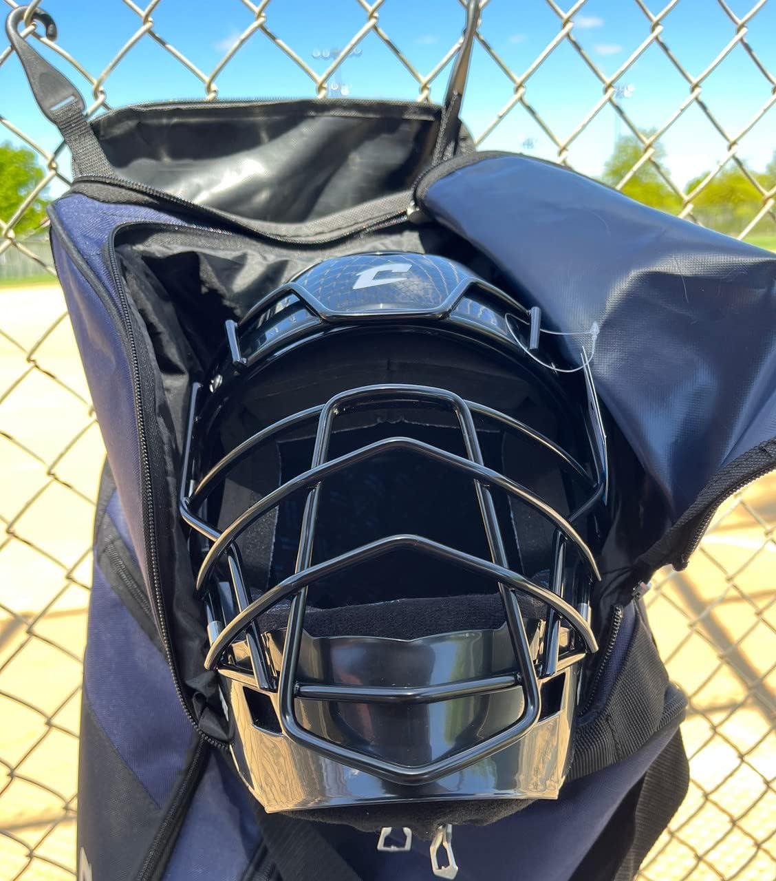 A Champro Boss Wheeled Catcher's Bag (E92) by Champro Sports hangs open on a chain-link fence, holding a black catcher's mask, with a baseball field and blue sky in the background.