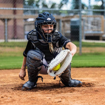 A softball catcher wearing the Mizuno Samurai Women's Catcher's Chest Protector (380407/380402) crouches behind home plate, reaching for a yellow softball on a dirt field, with a fence and trees in the background.