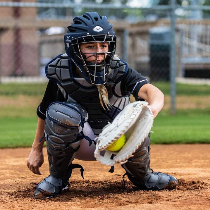 A softball catcher in full protective gear crouches behind home plate, glove extended to catch a yellow softball, during a game on an outdoor field.