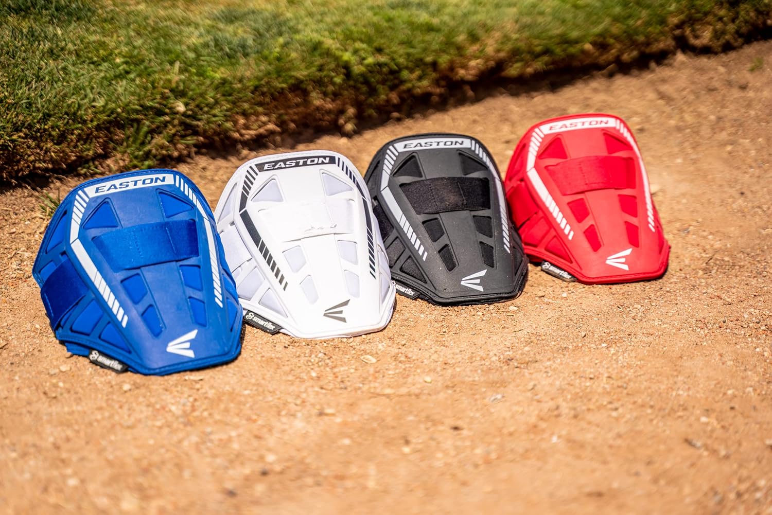 Four Easton chest protectors in blue, white, black, and red are lined up on dirt beside grass under sunlight, shown alongside the Easton Compact Batter Elbow Guard: ES37 for extra protection.