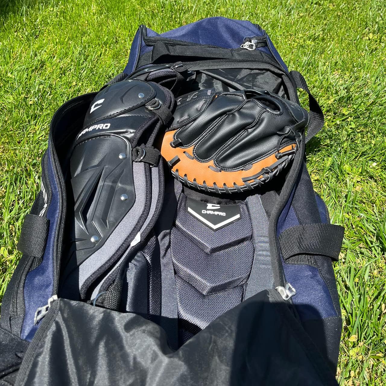A Champro Sports Champro Boss Wheeled Catcher's Bag: E92 in navy blue rests on grass, unzipped to display black and brown baseball gloves and black protective gear inside, with sunlight casting shadows on the equipment and bat pocket.