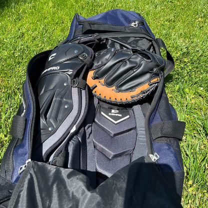 A Champro Sports Champro Boss Wheeled Catcher's Bag: E92 in navy blue rests on grass, unzipped to display black and brown baseball gloves and black protective gear inside, with sunlight casting shadows on the equipment and bat pocket.
