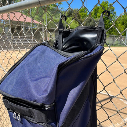 The Champro Sports Boss Wheeled Catcher's Bag (E92) in navy blue hangs open on a chain-link fence at a sunny baseball field, showing its spacious bat pocket with the diamond and dugout visible in the background.